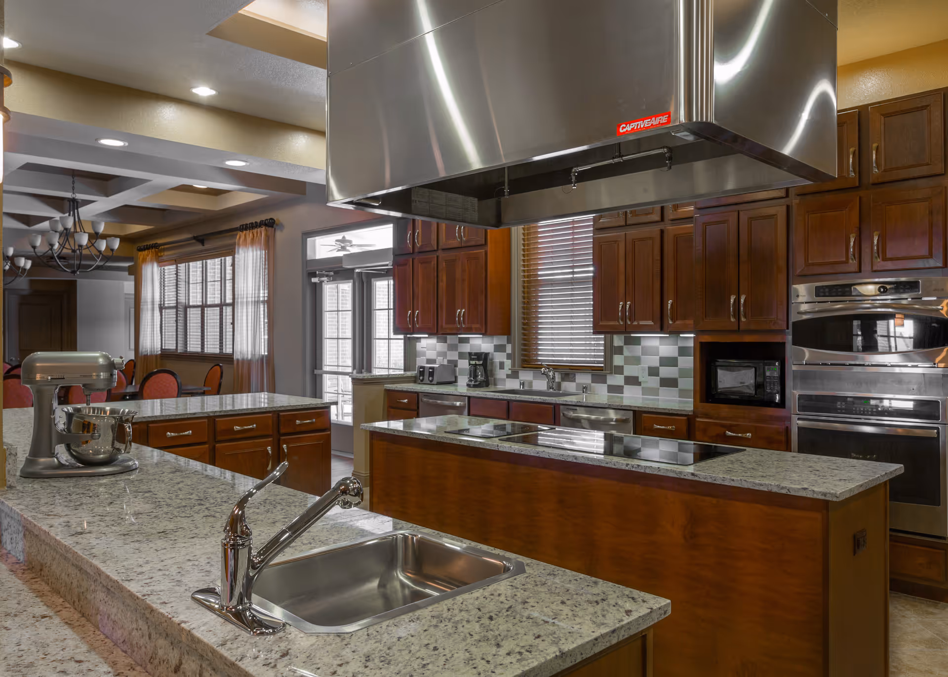 Communal kitchen with granite islands, a stainless-steel hood and appliances, and dark wooden cabinets.