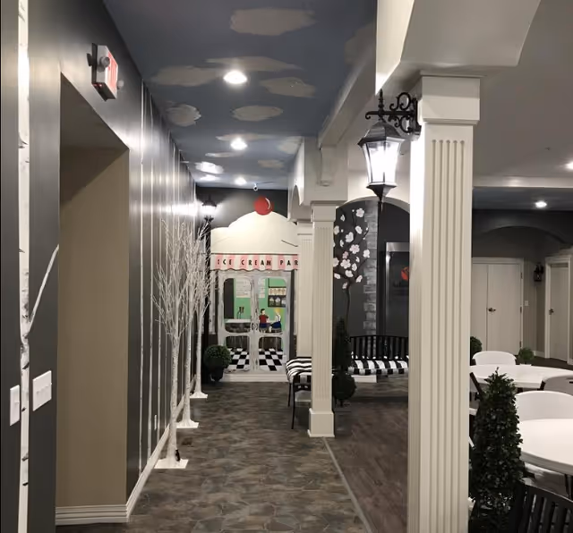 Interior hallway of a senior living facility decorated with white tree murals on the left wall and white columns on the right. The ceiling is painted blue with white clouds. At the end of the hallway, there is a mural of an ice cream parlor with a red cherry on top. To the right, there are black and white striped benches and white tables with chairs, along with small green plants. The space is well-lit with ceiling lights and wall-mounted lantern-style lamps.