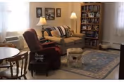 A cozy living room with a beige couch, a dark recliner chair, a round wooden table with chairs, two table lamps, a bookshelf filled with books, framed pictures on the wall, and a patterned area rug on the floor.