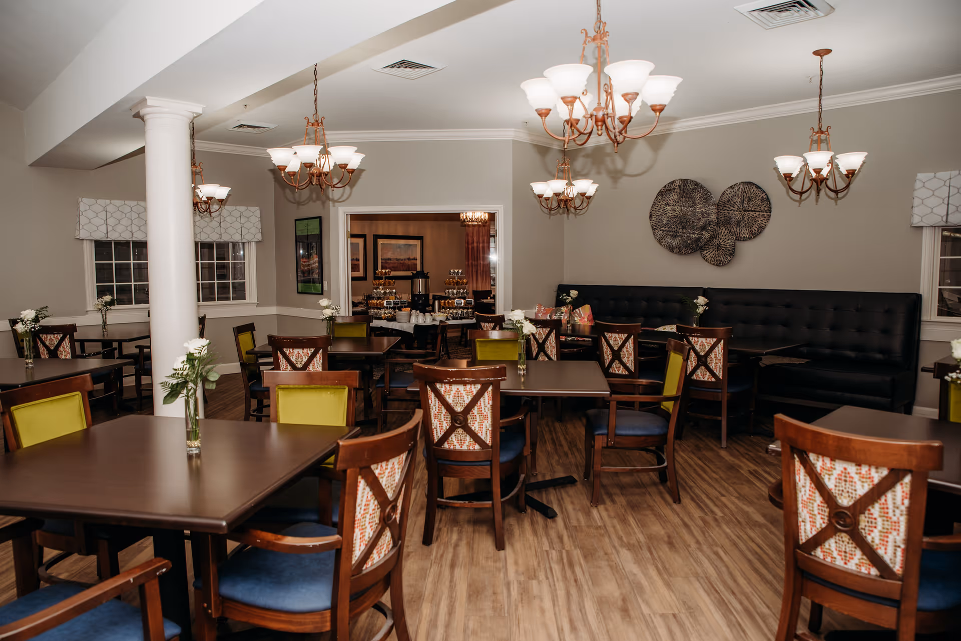 A dining room with multiple wooden tables and chairs, some with green cushions and others with patterned backs. The room has wooden flooring, beige walls, and several chandeliers hanging from the ceiling. There are windows with patterned valances and decorative wall art. In the background, there is a buffet table with food and beverages.