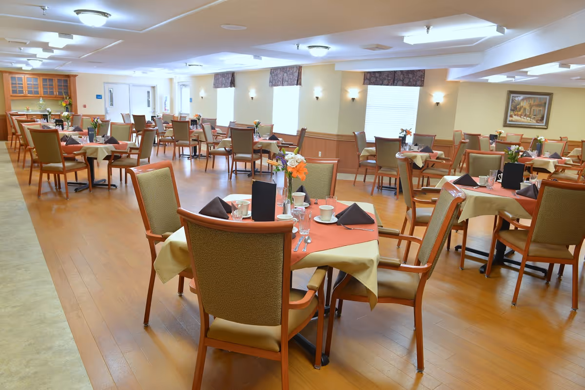 A spacious dining room with multiple tables covered with beige and orange tablecloths, each set with glasses, cups, silverware, and napkins. The room has wooden flooring, beige walls with wall sconces, and several windows with valances. There are wooden chairs with cushioned seats around each table, and a painting is visible on the far wall.