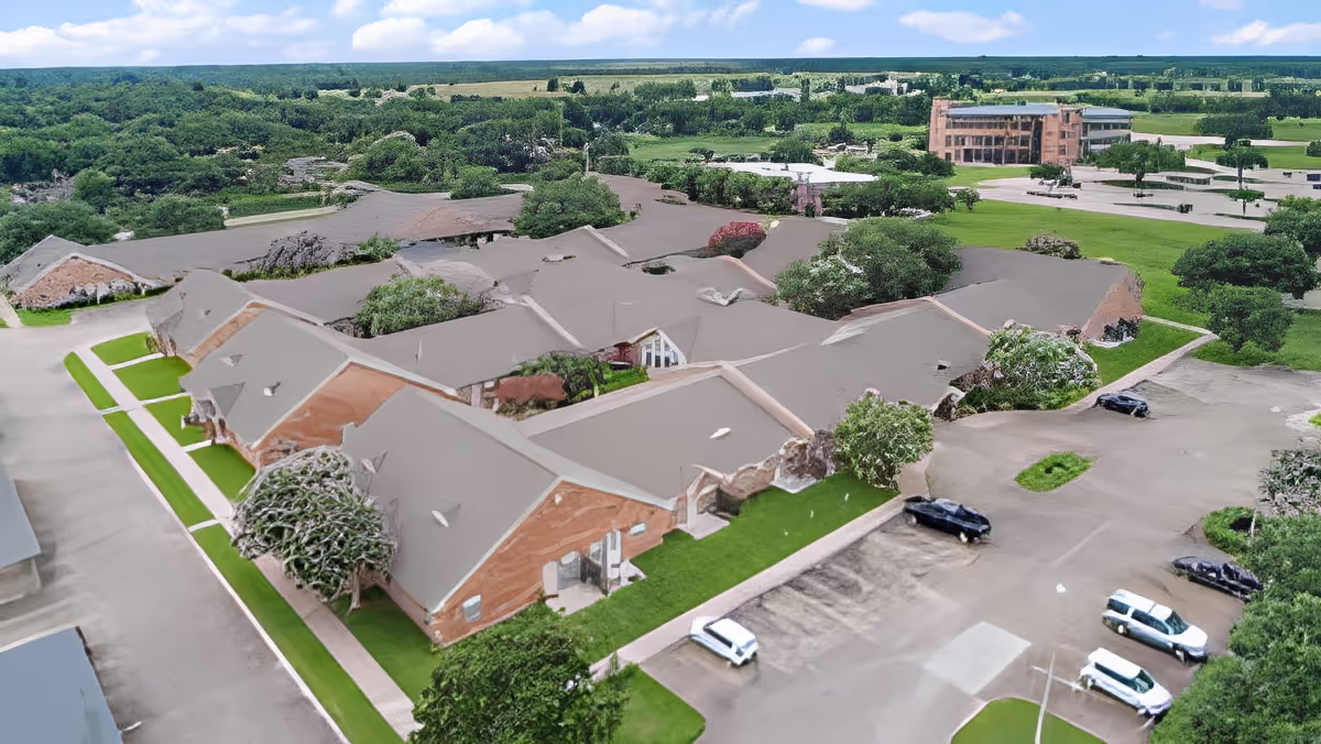 Aerial view of Morada Victoria senior living facility showing multiple connected brick buildings with gray roofs surrounded by green lawns, trees, and parking lots with several parked cars under a partly cloudy sky.