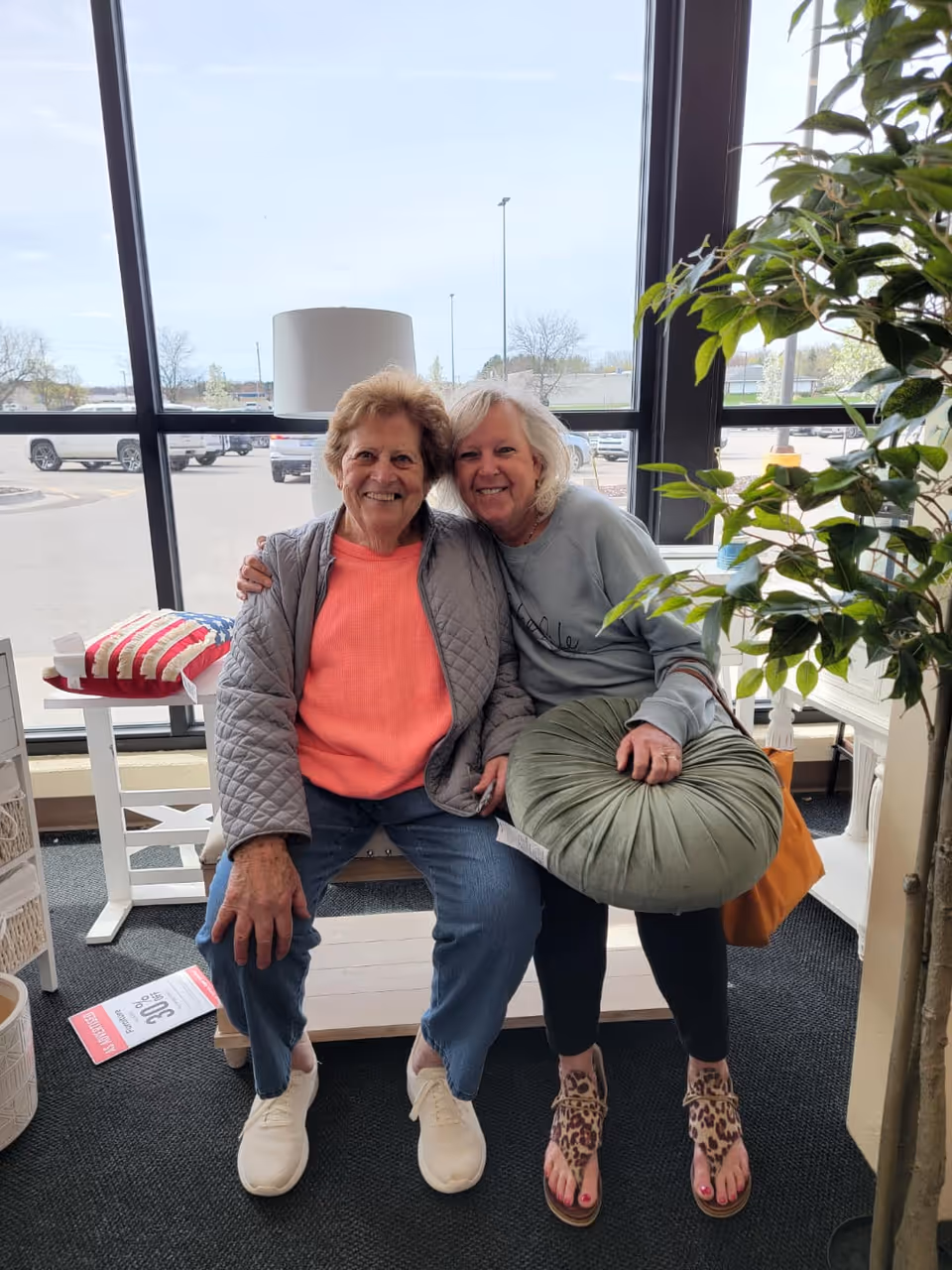 Two elderly women sitting closely together on a wooden bench inside a room with large windows showing a parking lot outside. One woman is wearing a coral sweater and gray jacket, and the other is wearing a light gray sweatshirt and holding a round green cushion. There is a lamp and a small table with a red, white, and blue pillow behind them, and a potted plant is visible on the right side.