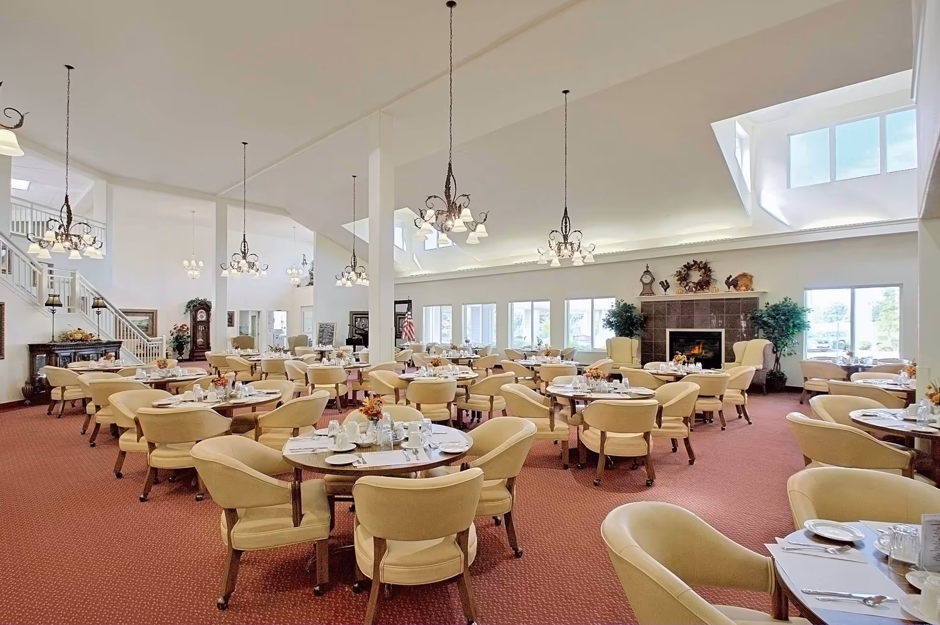 A spacious dining room with multiple round tables set with white plates, cups, and silverware. The room features beige cushioned chairs, red carpet flooring, large windows allowing natural light, and a fireplace with decorative items on the mantel. Several chandeliers hang from the high ceiling, and there is a staircase and grandfather clock visible in the background.