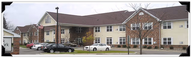 Exterior view of a two-story senior living facility building with beige siding and brick accents. Several cars are parked in front of the building, and a flagpole with an American flag is visible near the entrance. Trees without leaves are in the foreground.