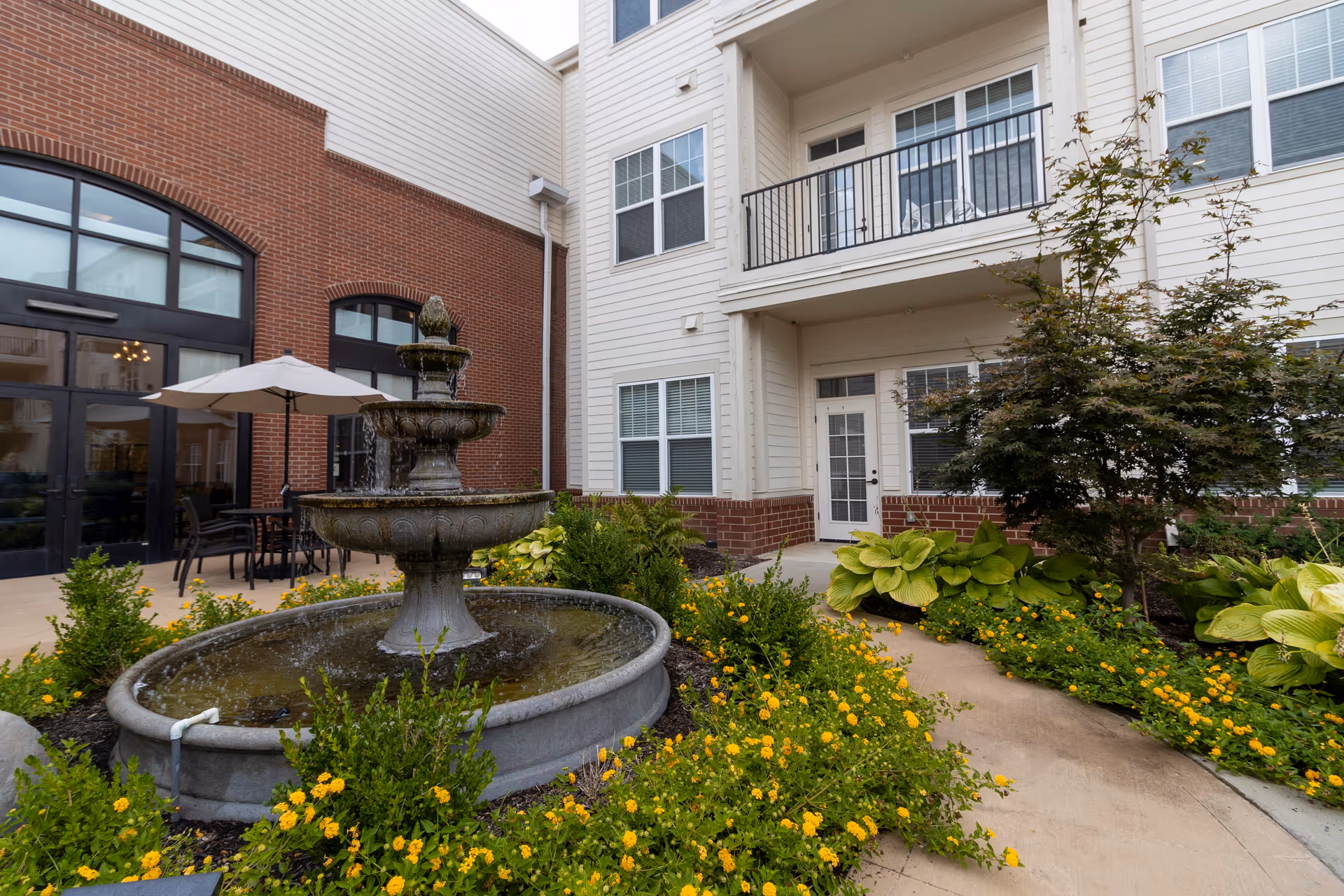 Outdoor courtyard area of a senior living facility featuring a three-tiered water fountain surrounded by yellow flowers and green shrubs. The courtyard is bordered by a building with white siding and brick accents, with windows and a balcony visible. There is also a patio area with a table, chairs, and an umbrella near large glass doors.
