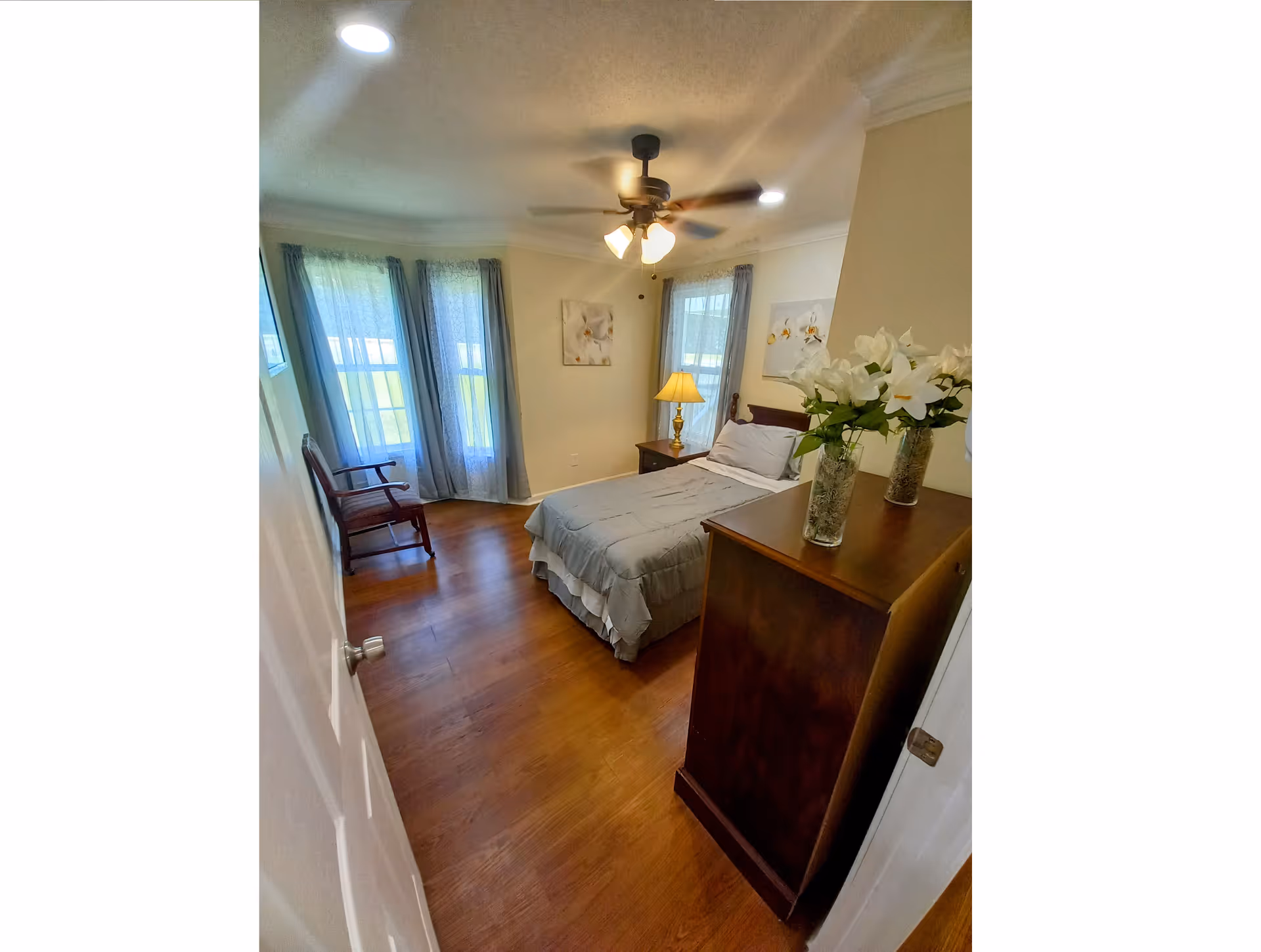Furnished bedroom with a single bed, wooden dresser topped with vases of flowers, a chair by tall windows with curtains, a ceiling fan, and hardwood floors.