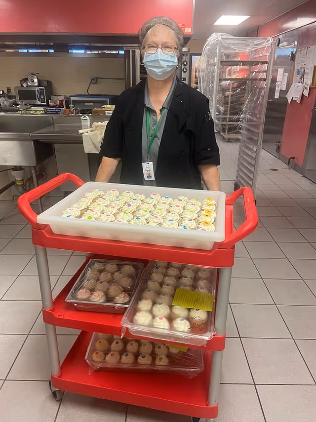 A masked staff member wearing a hairnet stands behind a red service cart loaded with trays of decorated cupcakes in a commercial kitchen.