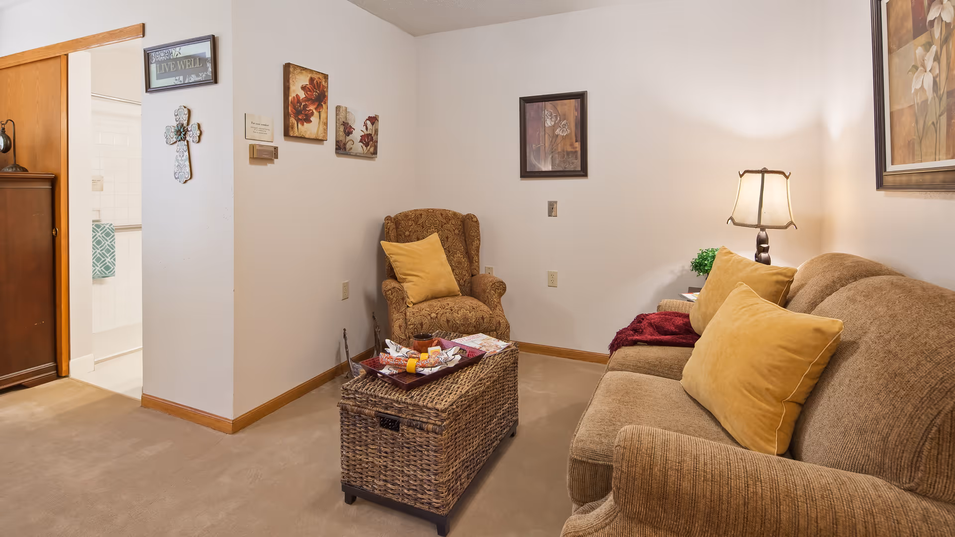 A cozy living room area with a brown upholstered sofa and matching armchair, both with yellow cushions. A wicker coffee table sits in front of the sofa with a tray of snacks on top. The walls are decorated with framed floral artwork and a cross. A lamp on a side table provides warm lighting. An open doorway leads to a bathroom with a towel hanging on the wall.