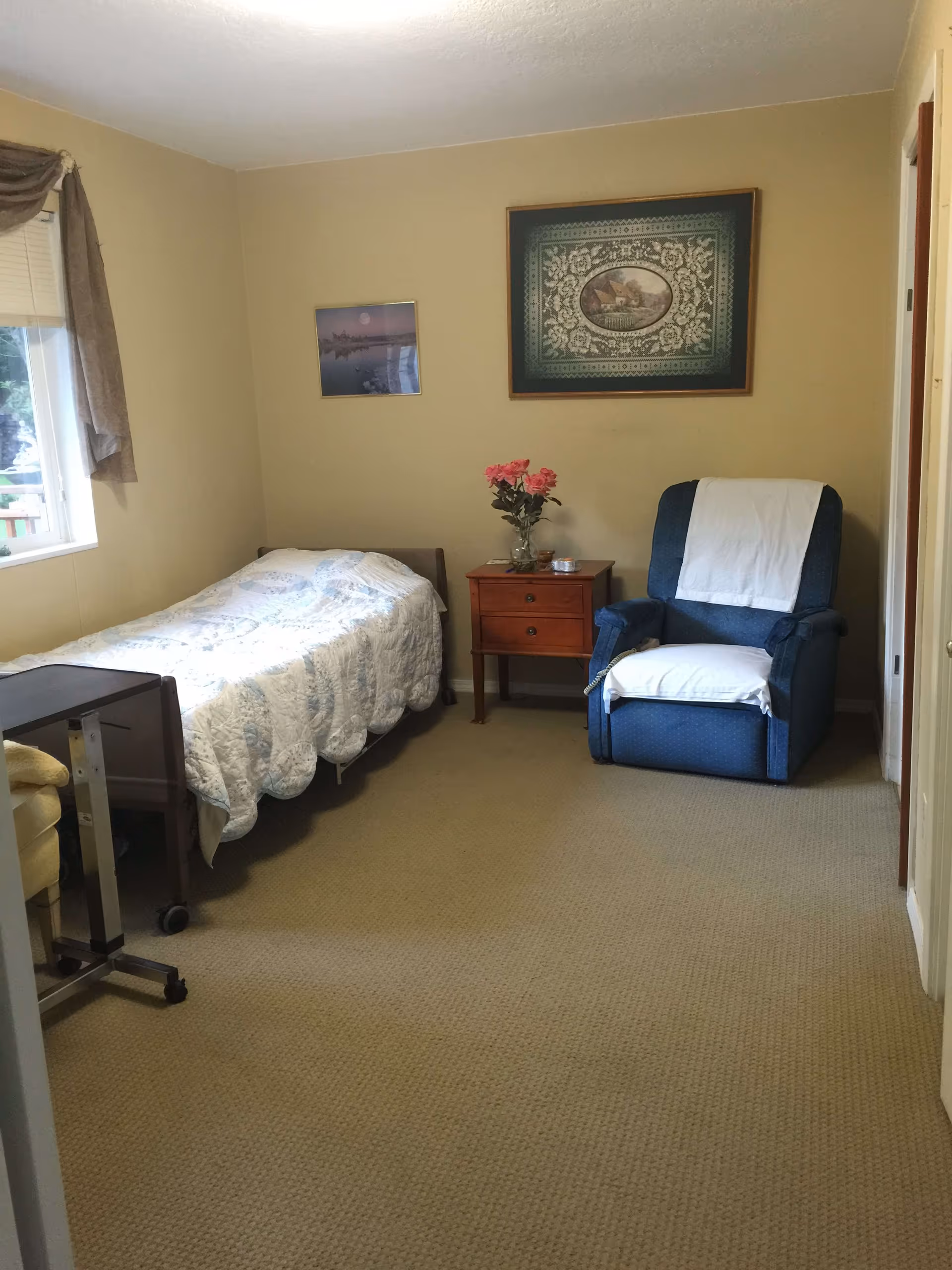 A small bedroom with a single bed covered with a white and blue quilt, a wooden nightstand with a vase of pink flowers, and a blue armchair with white covers on the headrest and seat. The walls are beige with two framed pictures hanging above the nightstand. A window with a brown curtain is on the left side of the room.