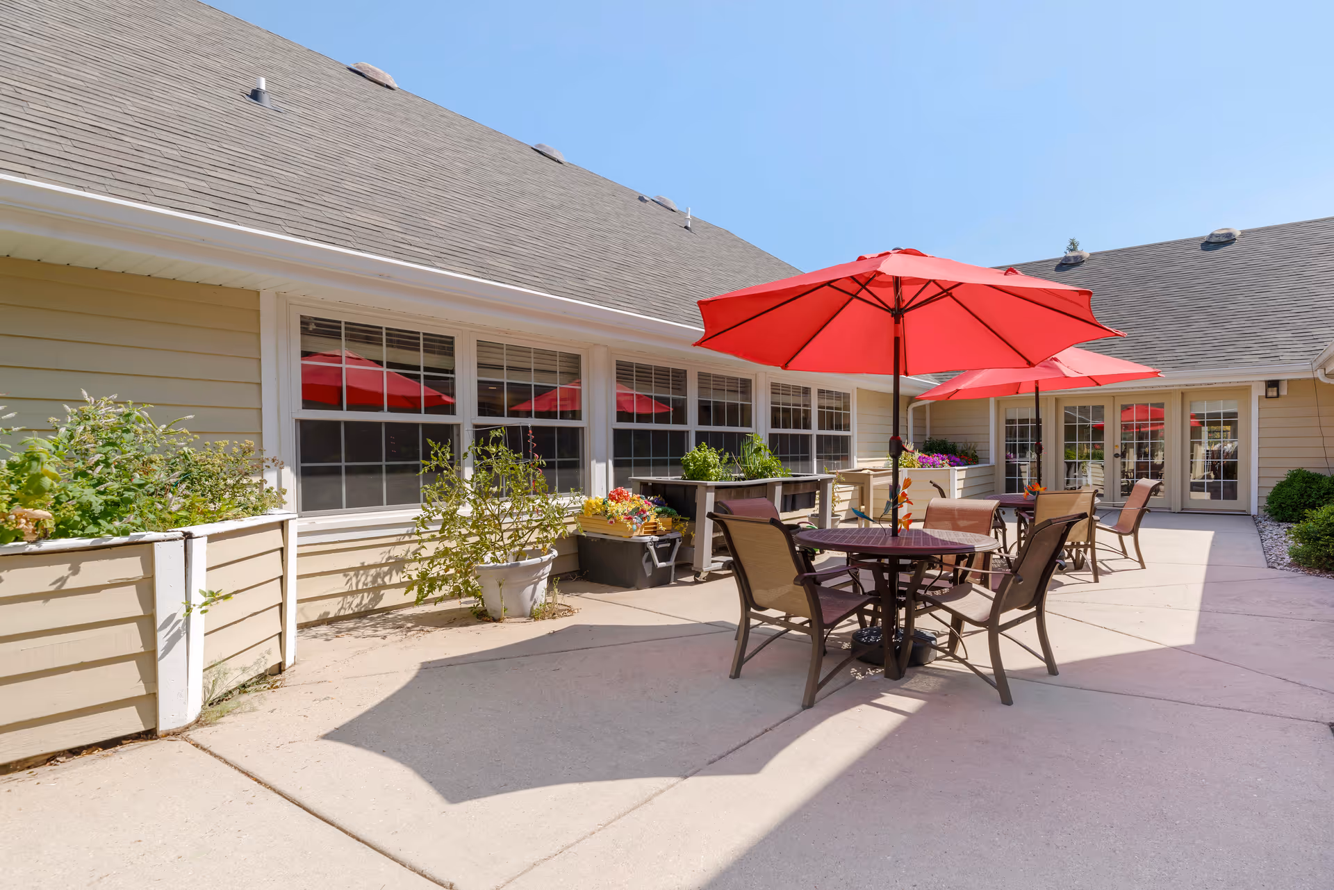 Outdoor patio area at Brookdale Brookfield Capitol Dr with several round tables and chairs under red umbrellas. Raised garden beds with plants and flowers are placed along the building wall. The building has beige siding and multiple windows, with a clear blue sky overhead.