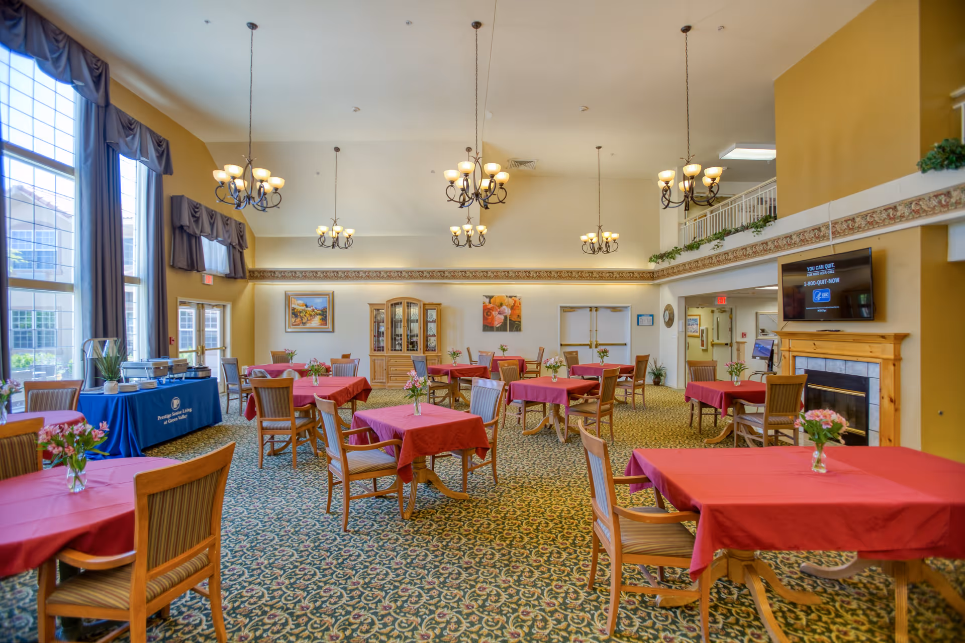 A spacious dining room with multiple tables covered in red tablecloths, each adorned with a small vase of flowers. Wooden chairs surround the tables. Large windows with dark curtains allow natural light to fill the room. Several chandeliers hang from the high ceiling. A fireplace with a TV mounted above it is visible on the right side. A buffet table covered with a blue cloth is set up near the windows.