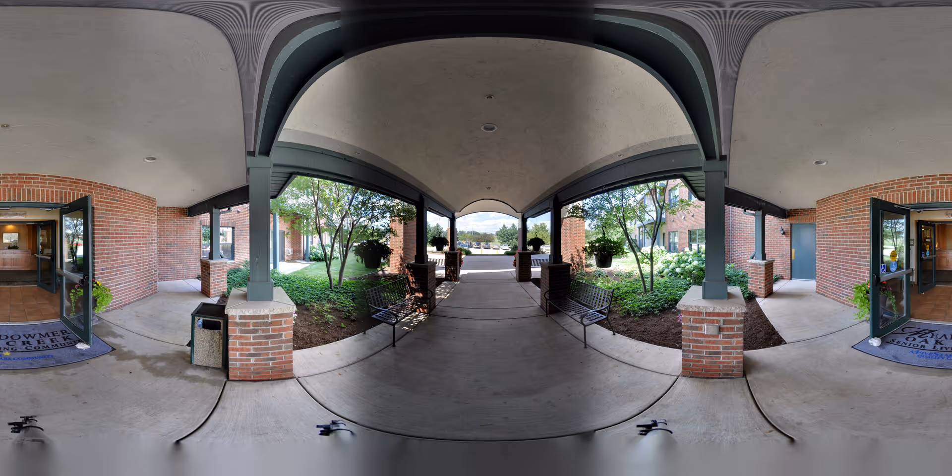 Covered brick entrance with columns, benches, landscaping, and double glass doors leading into a senior living facility.