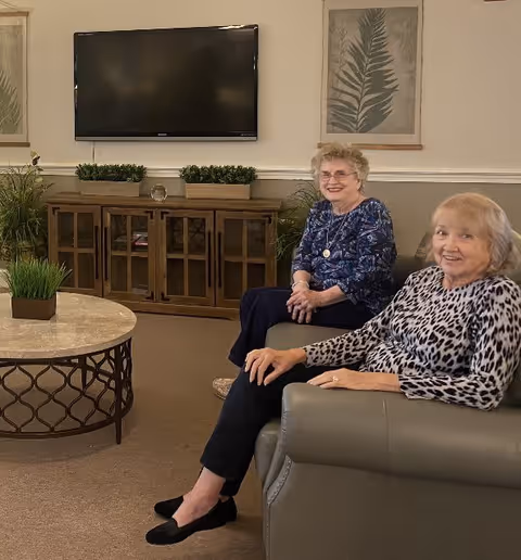 Two elderly women sitting and smiling in a living room area with a TV mounted on the wall, a wooden cabinet below the TV, a round coffee table with a small plant, and framed botanical artwork on the walls.