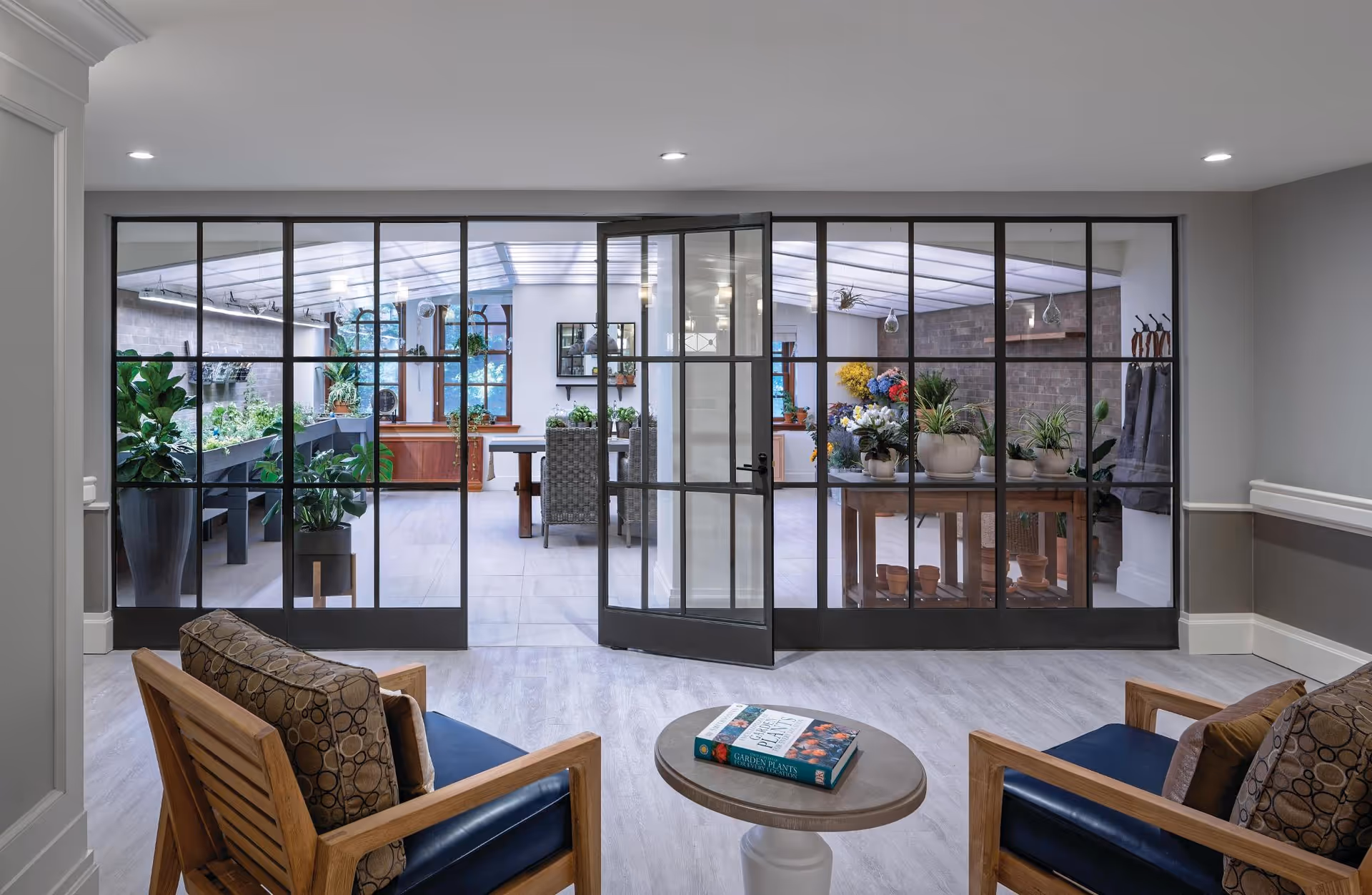 A cozy indoor seating area with two wooden armchairs featuring patterned cushions and a small round table with a book on gardening. Beyond the seating area, large glass doors with black frames open into a bright sunroom filled with various potted plants and flowers, a wooden table, and chairs. The space has a modern, clean design with light-colored flooring and walls.