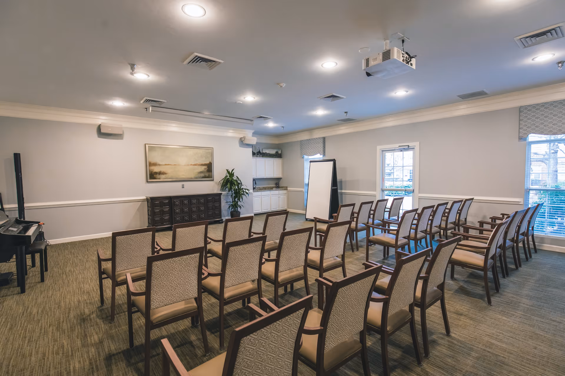 A well-lit meeting or activity room with rows of beige cushioned chairs arranged facing a whiteboard and a piano. The room has light gray walls, a carpeted floor, a large painting on the wall, windows with blinds, and a ceiling-mounted projector.