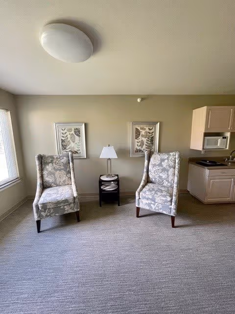 A small seating area with two floral patterned armchairs facing forward, separated by a small black side table with a white lamp on it. Behind the chairs are two framed botanical prints hanging on a beige wall. To the right is a kitchenette with light-colored cabinets, a microwave, and a sink. A window is partially visible on the left side, letting in natural light.