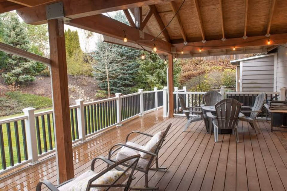 Covered wooden deck with chairs and a table under string lights overlooking a landscaped yard and trees.