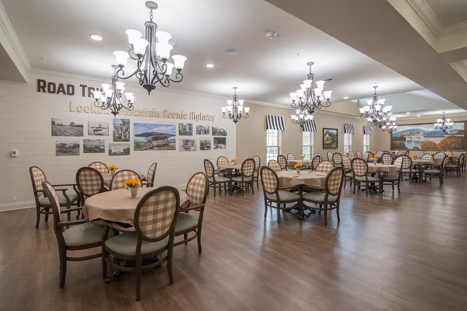 A spacious dining room with multiple round tables covered with beige tablecloths, each surrounded by four chairs with plaid upholstery. The room has wooden flooring, several chandeliers hanging from the ceiling, and windows with striped valances. The walls are decorated with framed pictures and a large mural, with text on one wall reading 'ROAD TRIP Lookout Mountain Scenic Highway.'