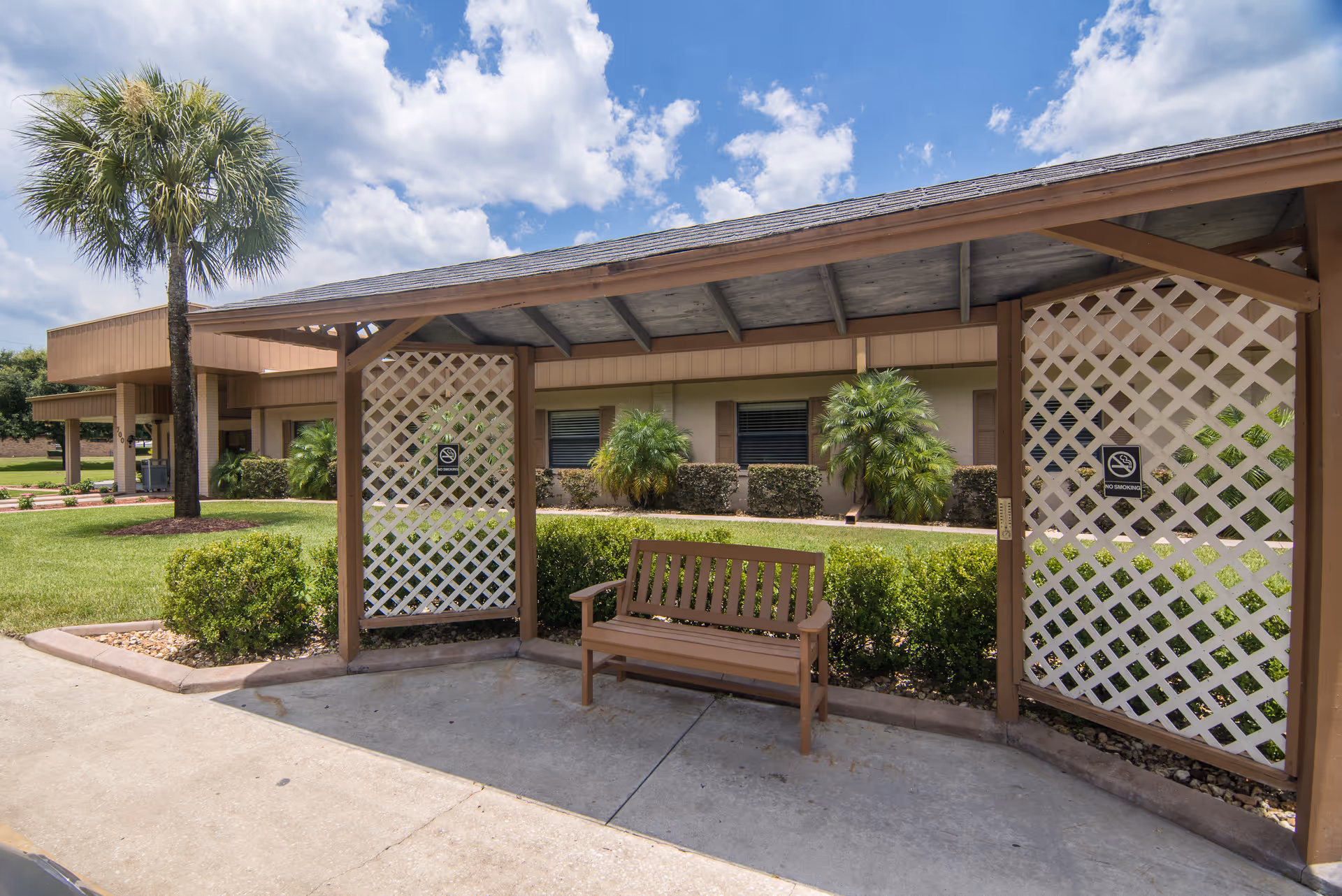 Outdoor covered seating area with a wooden bench, lattice panels on the sides, surrounded by green bushes and palm trees, with a building and partly cloudy sky in the background.