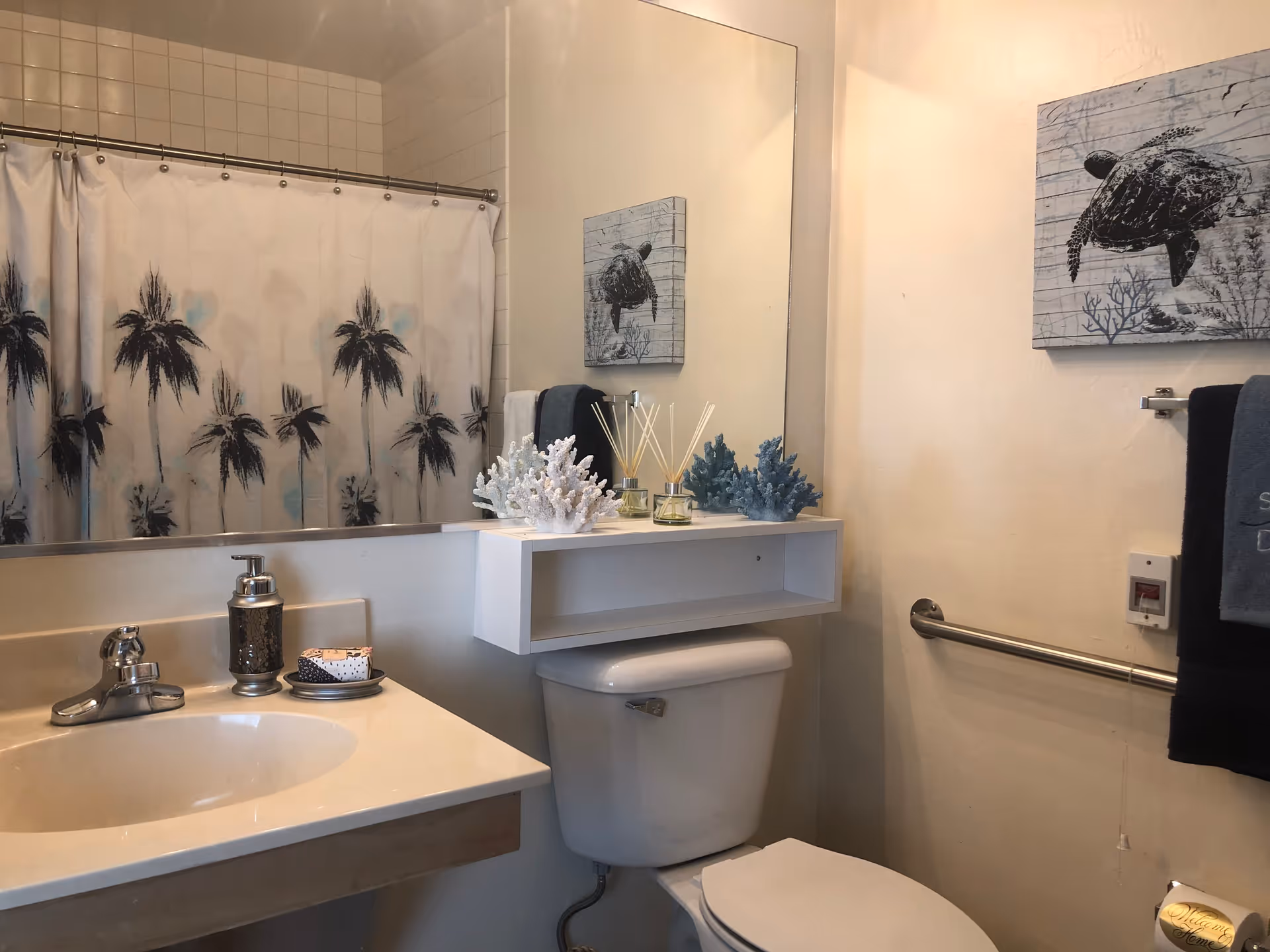 A bathroom with a white sink and countertop, a silver faucet, and a soap dispenser. Above the sink is a large mirror reflecting a shower curtain with a palm tree design. There is a white toilet beneath a white floating shelf holding decorative coral pieces and reed diffusers. On the wall next to the toilet is a towel rack with a dark towel and a framed picture of a sea turtle.