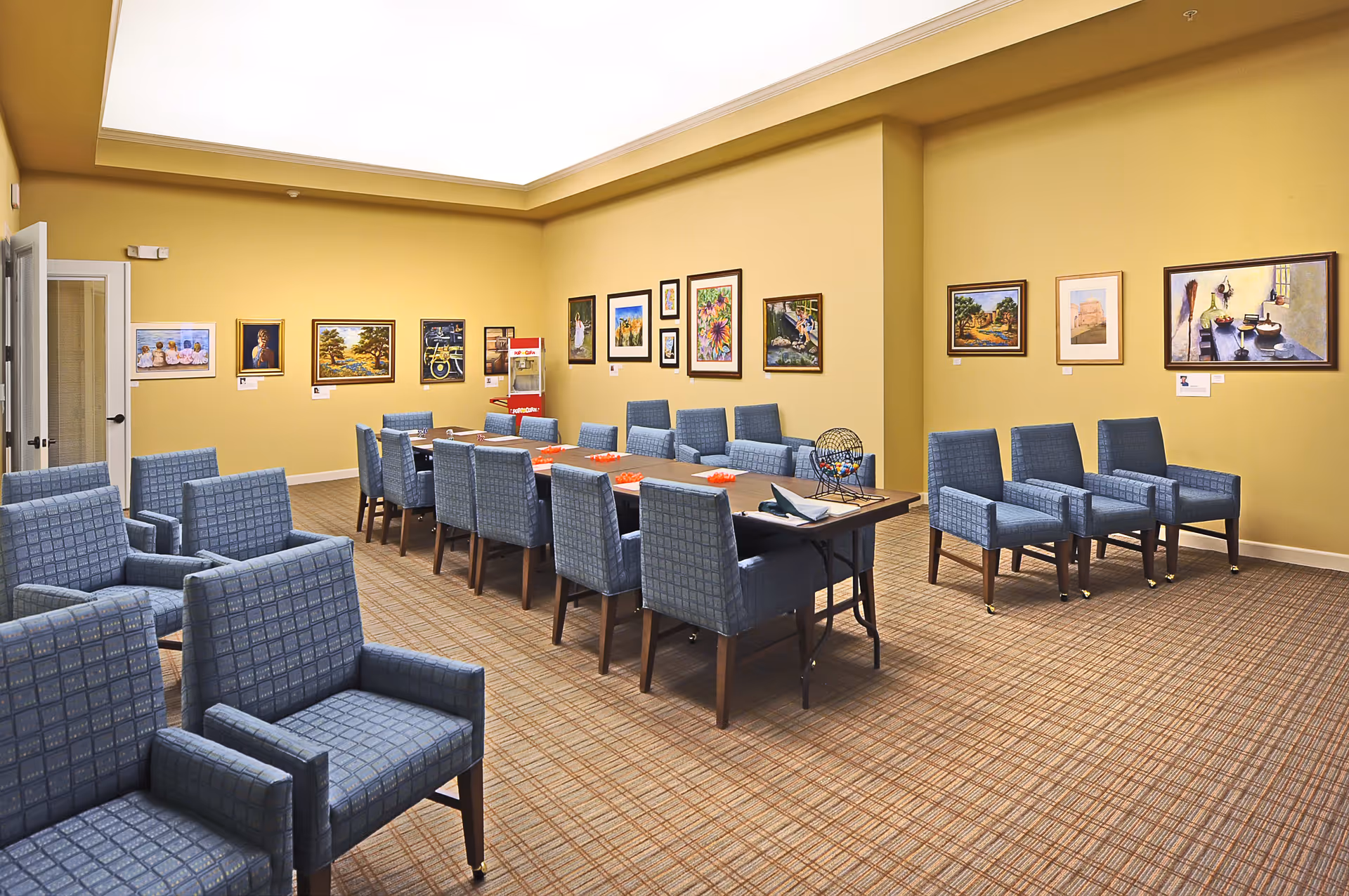 A well-lit room with yellow walls featuring multiple framed paintings. The room contains several blue upholstered chairs arranged around a long table and in rows along the walls. There is a bingo cage on the table, indicating the room is used for group activities or games.