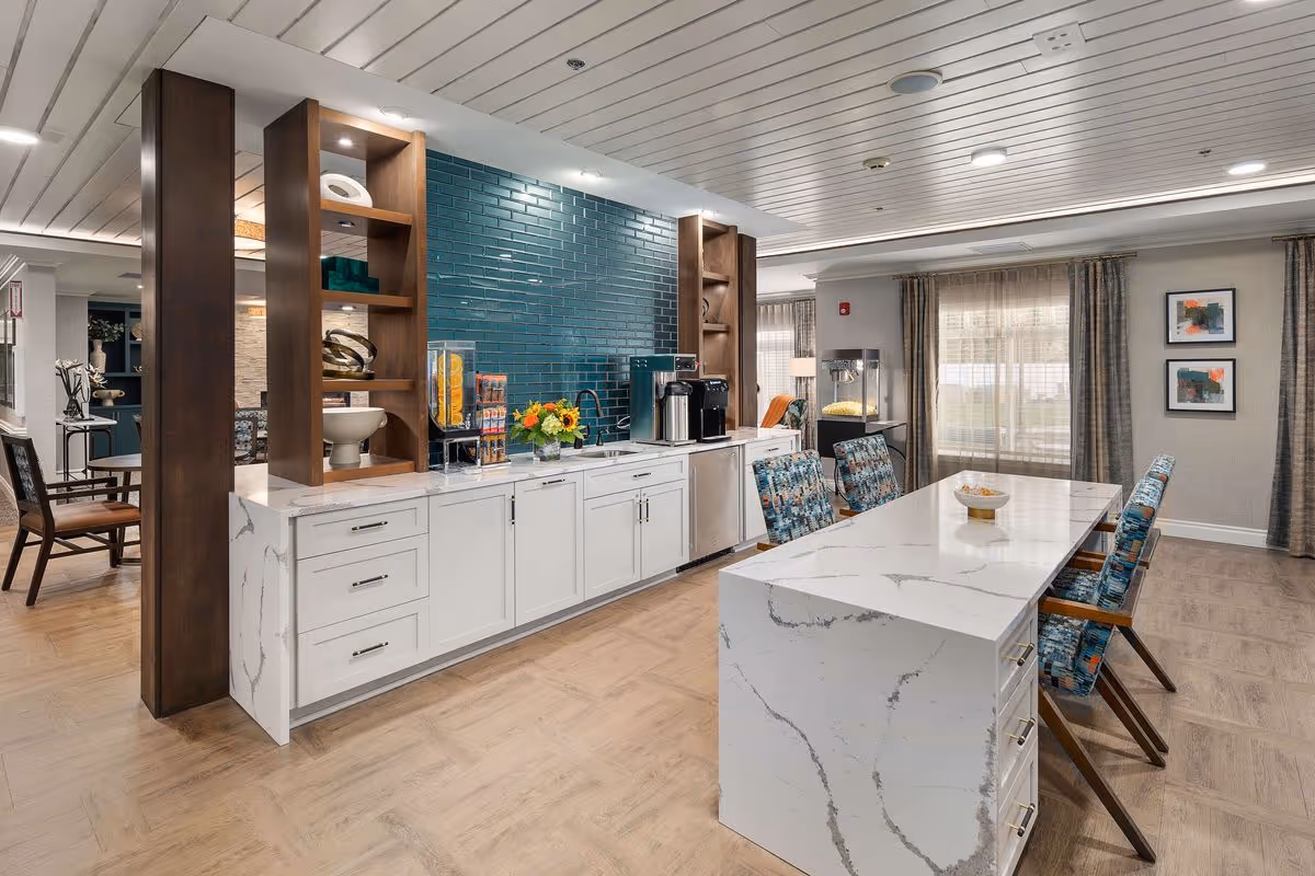 A modern communal kitchen and dining area with white marble countertops, a blue tiled backsplash, and wooden shelving. The space includes a coffee machine, a juice dispenser, and a popcorn machine. There are six patterned chairs around a large island table, and large windows with sheer curtains letting in natural light.