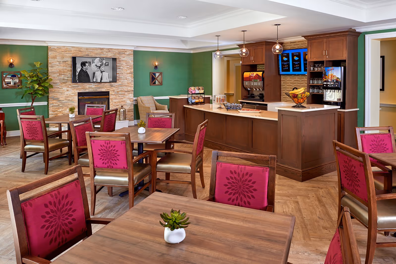 A dining area in an assisted living facility with wooden tables and chairs featuring red patterned upholstery. There is a counter with a beverage dispenser, fruit basket, and snacks. The walls are green with a stone fireplace and a TV mounted above it. Pendant lights hang above the counter, and there is a plant and decorative mirrors on the walls.