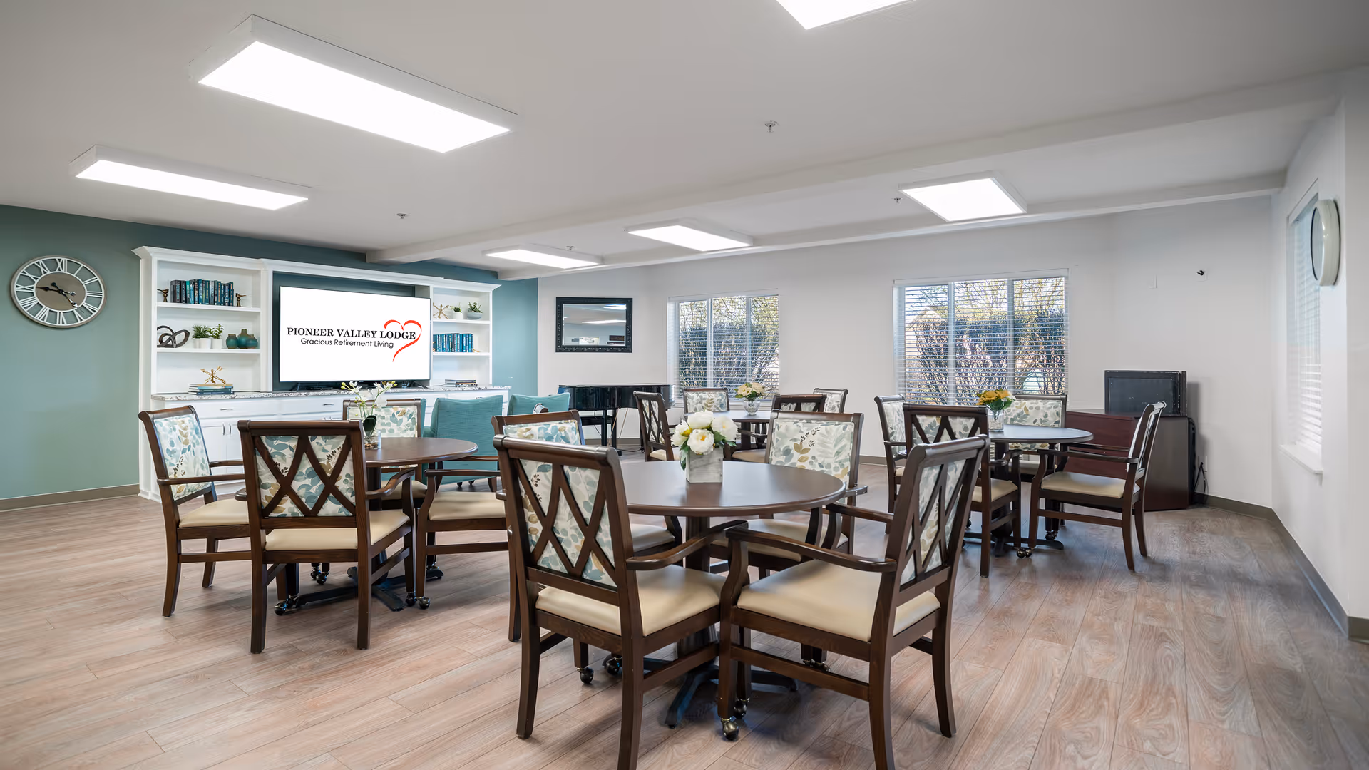 A bright and spacious common area in a retirement living facility with several round wooden tables surrounded by cushioned chairs featuring floral upholstery. The room has large windows with blinds, a white built-in shelving unit with decorative items and books, and a large screen displaying the text 'Pioneer Valley Lodge Gracious Retirement Living'. The walls are painted white and teal, and the floor has light wood-style flooring.