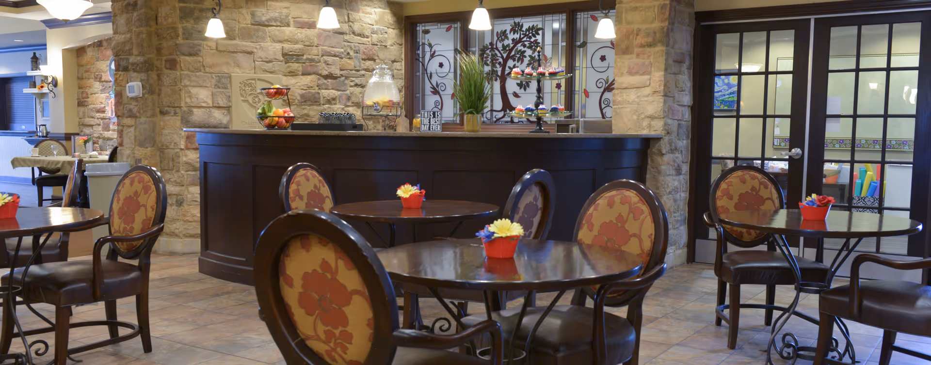 A cozy dining area with round wooden tables and chairs featuring floral upholstery. The room has stone walls and a dark wooden counter with decorative items including a fruit bowl and a tiered tray with cupcakes. Pendant lights hang from the ceiling, and there are glass doors with black frames in the background.
