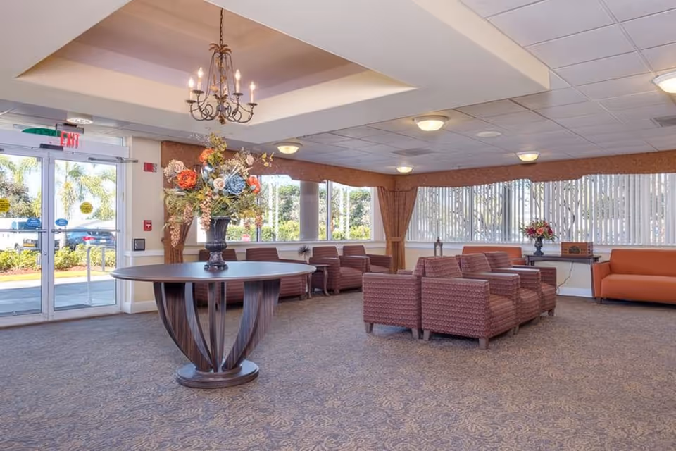 A spacious and well-lit living room area with multiple upholstered armchairs arranged in small groups, a large round wooden table with a floral arrangement in the center, large windows with vertical blinds, and an orange sofa along the wall. The room has a chandelier hanging from the ceiling and carpeted flooring.