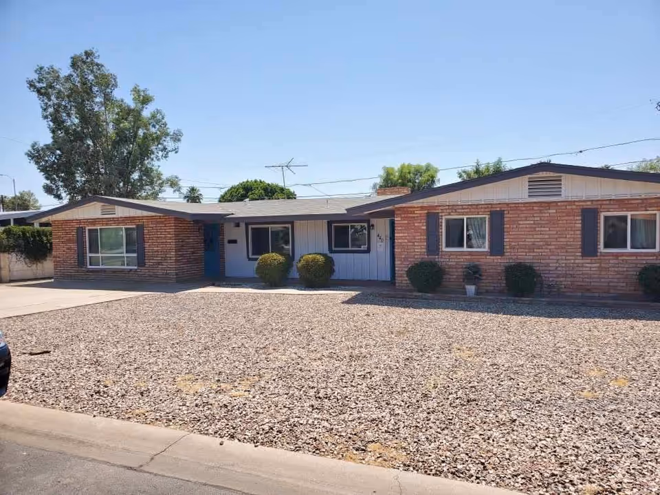 Single-story brick building with a gravel front yard and several bushes, under a clear blue sky.