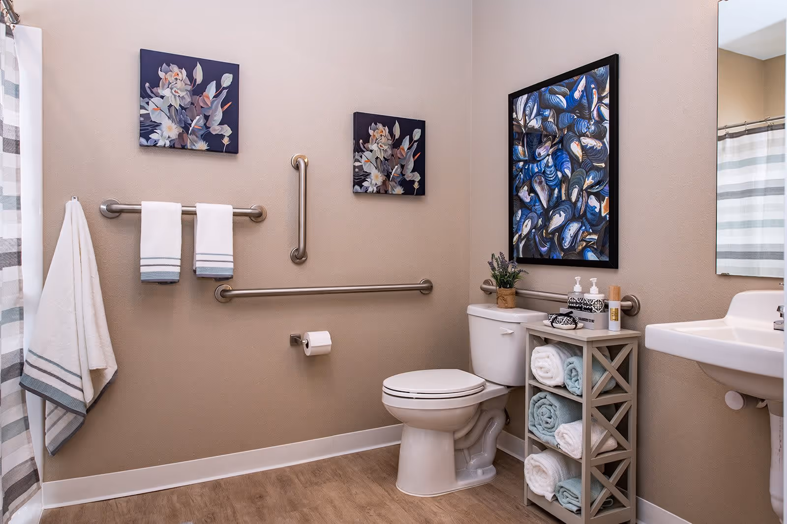 A clean and accessible bathroom with beige walls and wood-look flooring. The bathroom features a white toilet with a small shelf next to it holding rolled towels and toiletries. There are grab bars installed on the walls around the toilet and towel racks with white towels hanging. Two floral paintings are hung on the walls, and a shower curtain with gray and white stripes is partially visible. A white sink with a mirror above it is also visible.