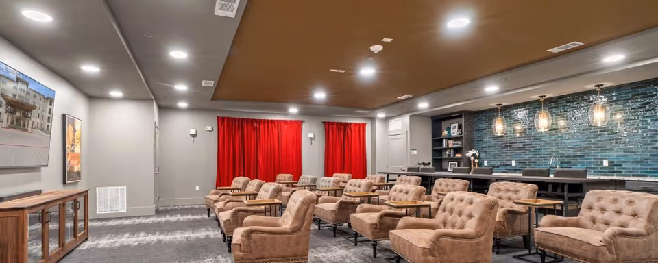 A spacious lounge area with multiple tan tufted armchairs arranged in rows, each with a small attached wooden table. The room features a dark brown ceiling, recessed lighting, and a wall with glossy teal subway tiles behind a long counter with bar stools. Red curtains cover the windows on the far wall, and a wooden cabinet with a large TV mounted above it is visible on the left side.