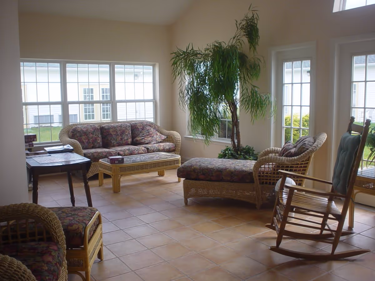 A bright living room with large windows letting in natural light. The room features wicker furniture with floral cushions, including a sofa, an armchair, a chaise lounge, and a coffee table. There is also a wooden rocking chair with a green cushion and a tall green potted plant near the windows. The floor is tiled with light brown tiles.