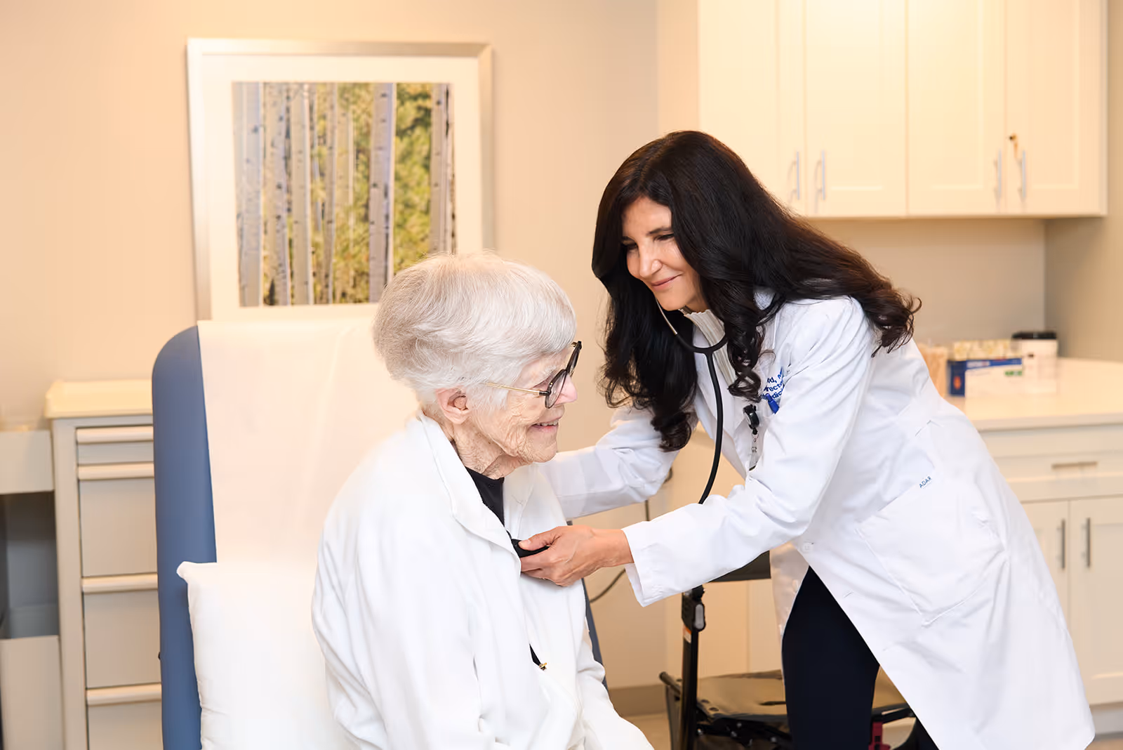 A female doctor with long dark hair uses a stethoscope to listen to the chest of an elderly woman with short white hair and glasses, who is seated in a medical examination chair in a clinical room with cabinets and a framed picture on the wall.