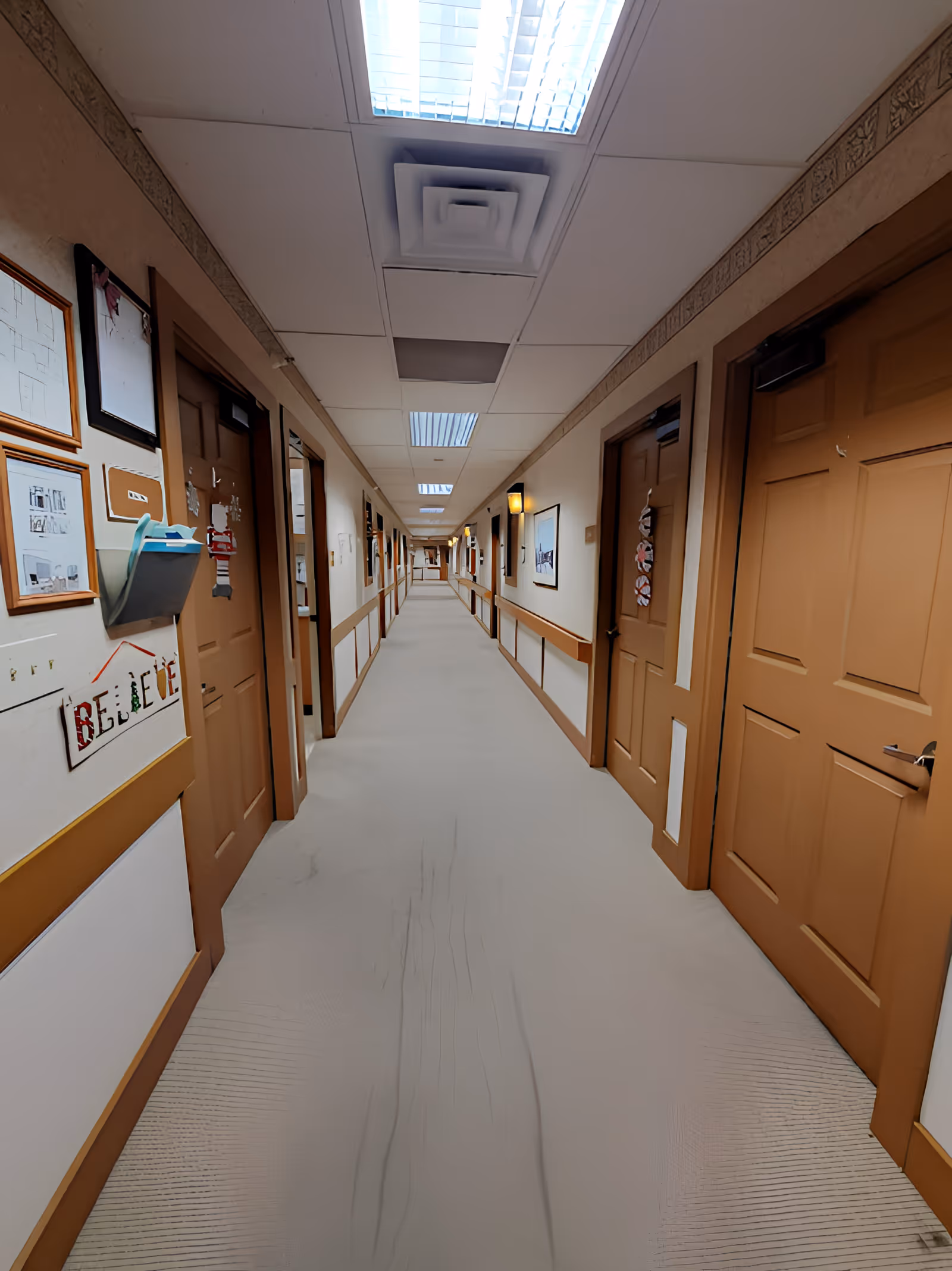 A long, well-lit hallway in a senior living facility with multiple closed wooden doors on both sides. The walls are decorated with framed pictures and a sign that says 'BELIEVE'. The ceiling has fluorescent lights and ventilation panels, and the floor is carpeted.