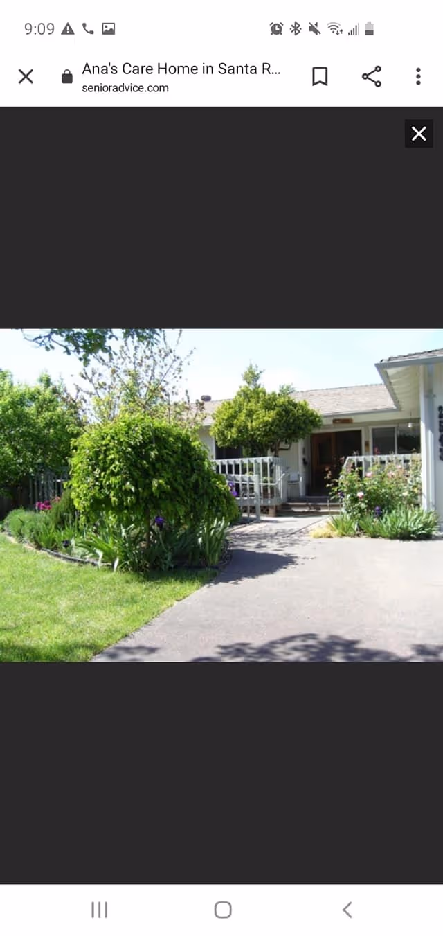 Front exterior view of a single-story care home building with a paved driveway, green lawn, and various shrubs and trees in the garden area.