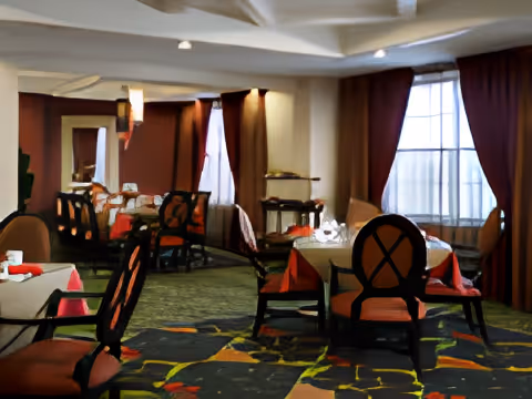 Interior view of a dining room with round tables covered with tablecloths and set with napkins and glassware. The room has large windows with curtains, cushioned chairs with dark wooden frames, and a patterned carpet.