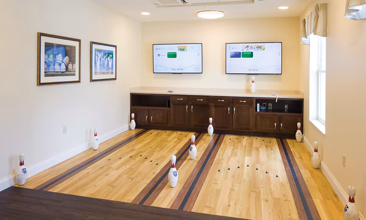 Indoor bowling alley with two lanes, wooden flooring, and bowling pins set up. Two flat-screen TVs are mounted on the wall above a wooden cabinet. The room has light-colored walls, two framed pictures on the left wall, and a window on the right side with curtains.
