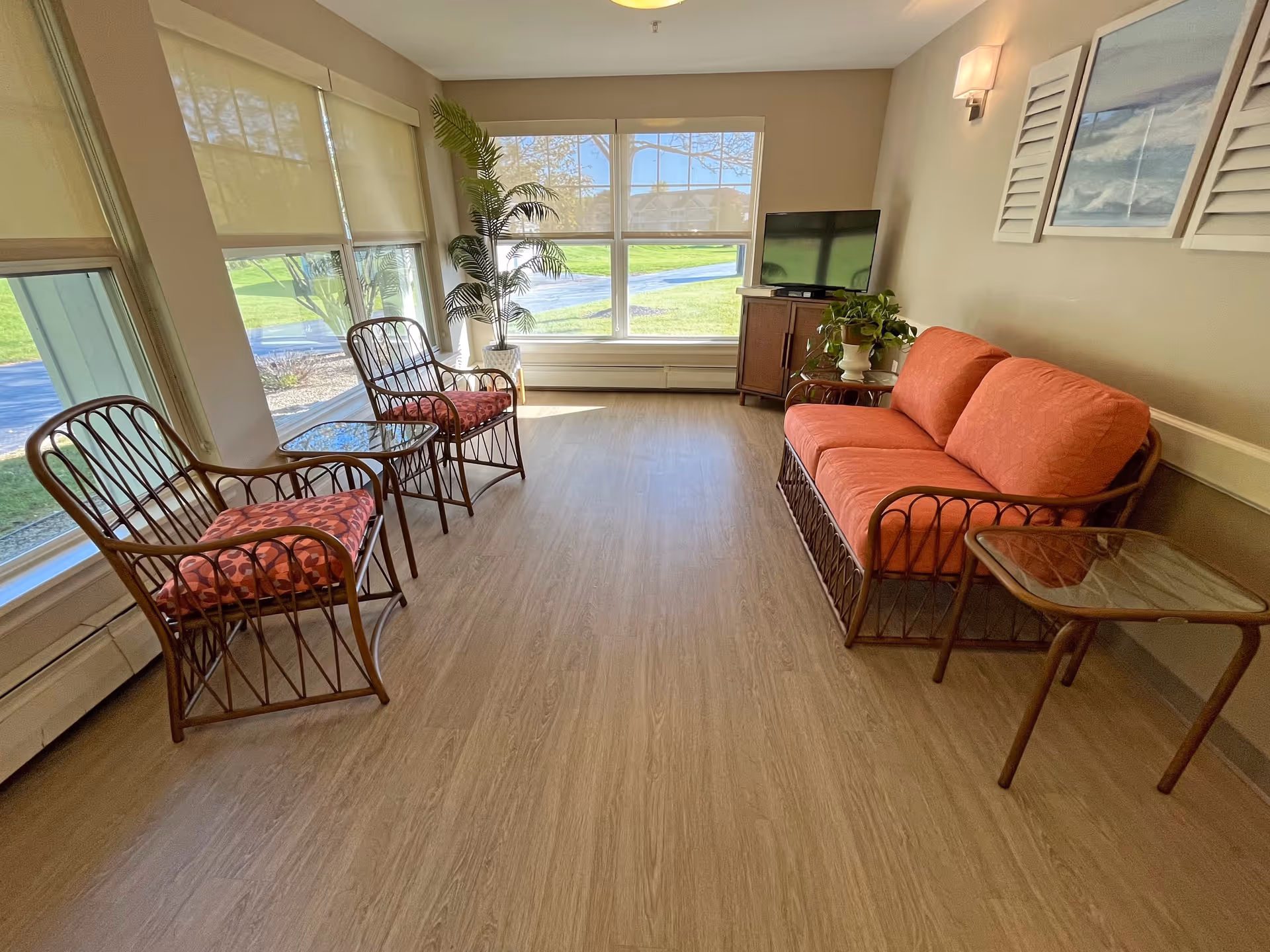 Sunlit sitting area with rattan sofa and chairs arranged by large windows overlooking a green lawn.