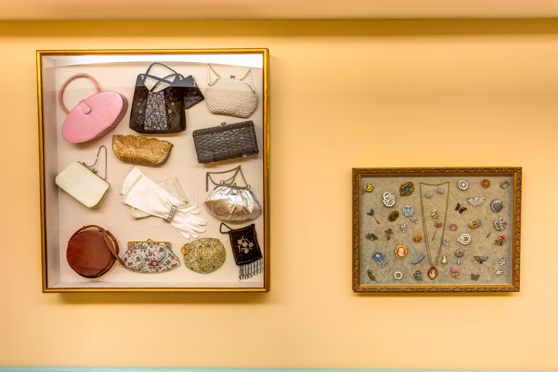 Two framed displays mounted on a beige wall. The left frame contains a collection of vintage handbags and a pair of white gloves. The right frame showcases an assortment of vintage brooches, pins, and necklaces arranged on a fabric background.