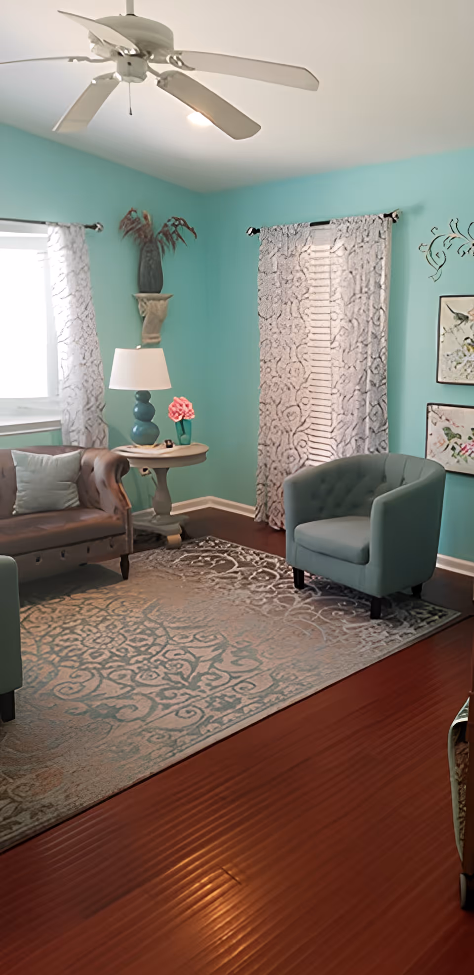 A cozy living room with turquoise walls, a ceiling fan, a brown leather sofa with a light blue pillow, a round side table with a lamp and a small flower vase, a light blue armchair, patterned curtains on two windows, decorative wall art, and a large patterned area rug on a wooden floor.