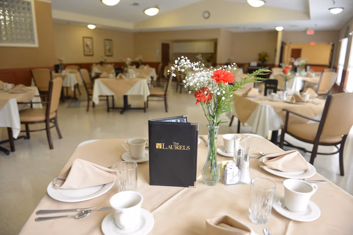 A dining room table set with plates, cups, a vase of flowers and a menu labeled 'The Laurels' with several other set tables in the background.
