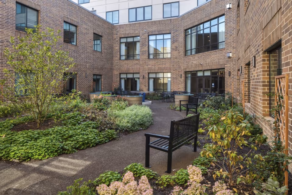 A peaceful outdoor courtyard area surrounded by a brick building with multiple windows. The courtyard features a paved walkway, green shrubs, flowering plants, small trees, and several black benches for seating.
