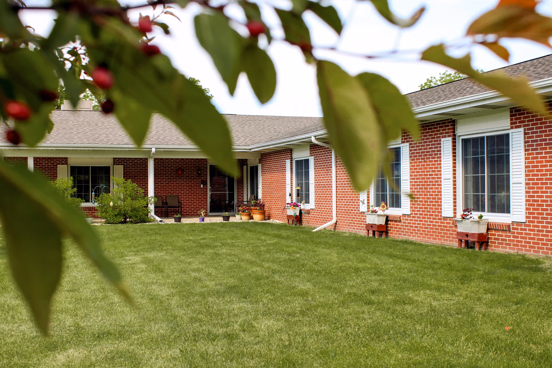 Single-story red brick senior living building with windows, flower boxes and a grassy lawn framed by tree leaves.