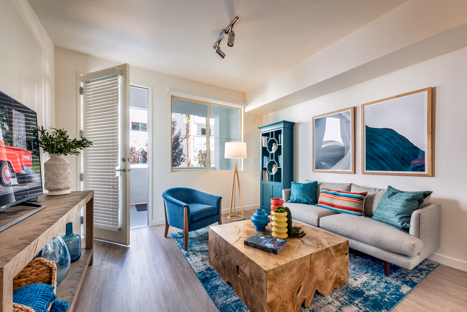 A bright living room with a gray sofa adorned with colorful pillows, a wooden coffee table with decorative items, a blue armchair, a tall floor lamp, a blue cabinet, and two framed abstract paintings on the wall. A wooden console table with a TV and decorative vases is on the left, and a door with blinds is open to a small outdoor area.