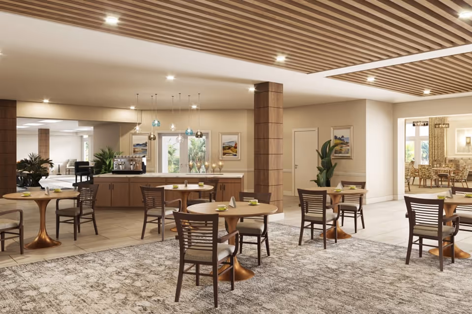 A spacious and well-lit dining area with round wooden tables and chairs arranged on a patterned carpet. The room features a wooden slatted ceiling design with recessed lighting. In the background, there is a counter with a coffee machine and pendant lights hanging above it. The walls are decorated with framed artwork and there are plants placed around the room. Adjacent to this area is another dining space with more tables and chairs visible through an open doorway.