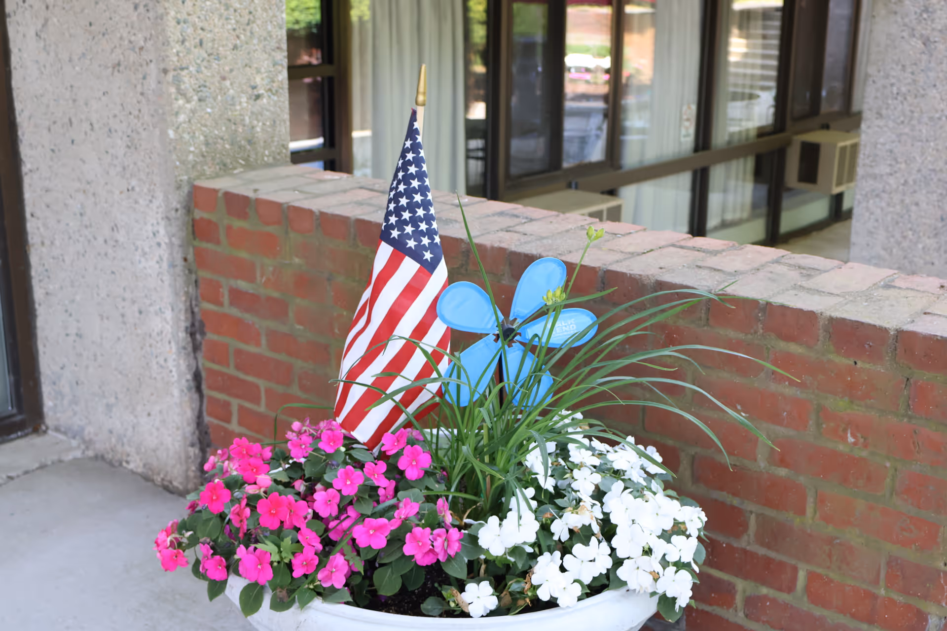 A white planter filled with pink and white flowers, green grass-like plants, an American flag, and a blue pinwheel shaped like a flower, placed on a concrete surface next to a brick wall and a building with large windows.