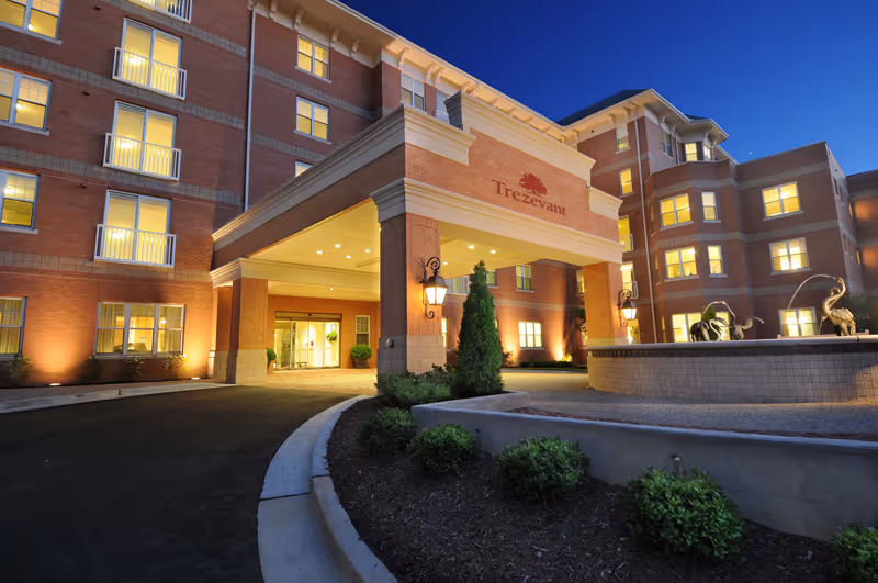 Exterior view of the Trezevant senior living facility at dusk, showing a multi-story brick building with illuminated windows and entrance canopy. There is a curved driveway leading to the entrance, landscaped greenery, and a decorative fountain with sculptures in front.
